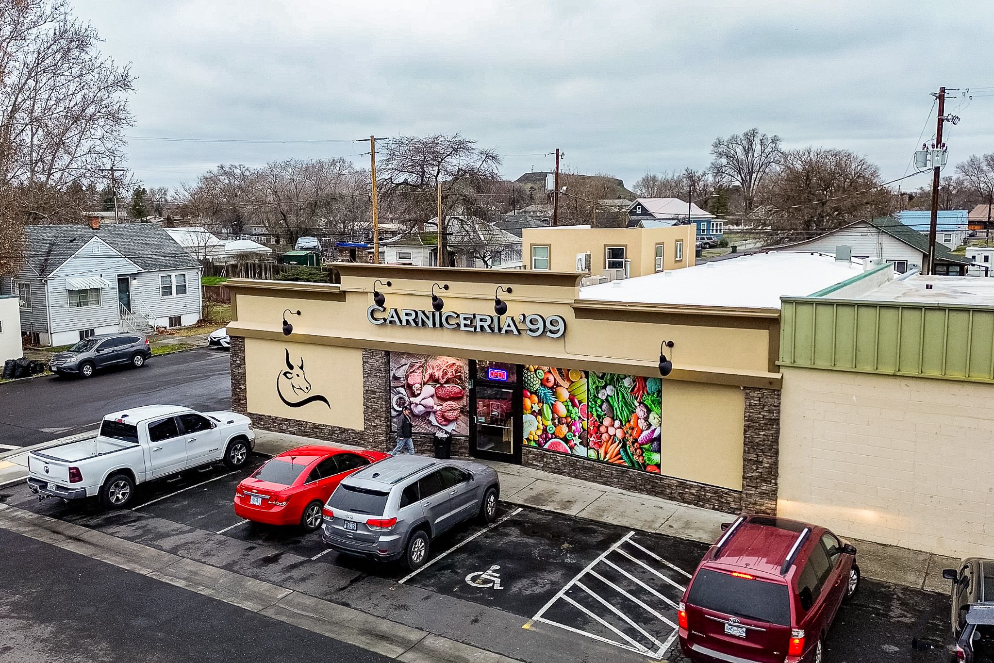 Aerial view of the Carnicería 99 market and parking lot in Hermiston, Oregon, with the bull logo and produce mural on the storefront