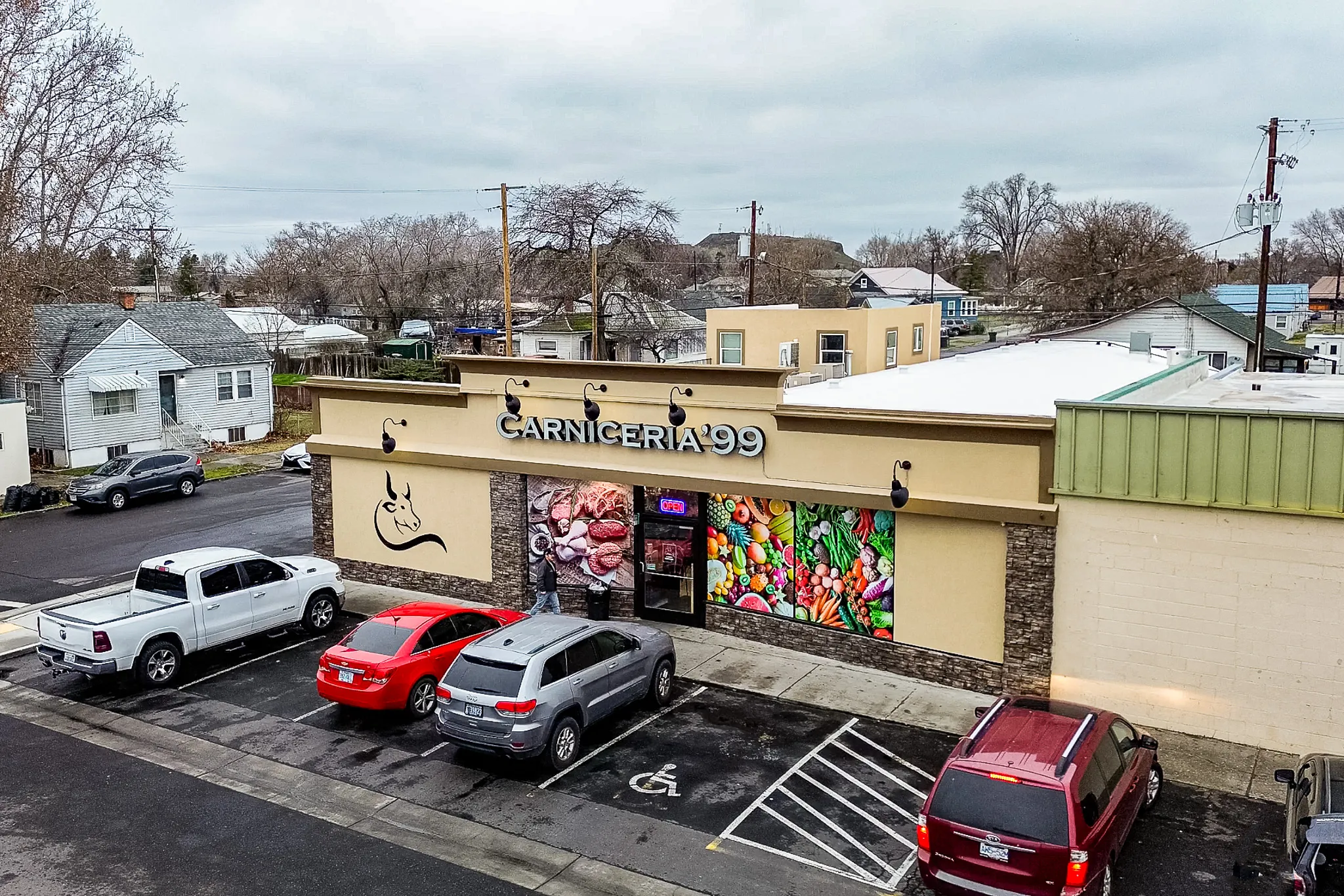 Aerial view of the Carnicería 99 market and parking lot in Hermiston, Oregon, with the bull logo and produce mural on the storefront